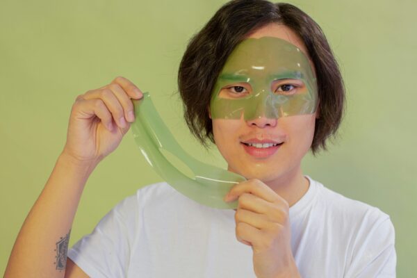 Asian man applying a green facial mask for skincare in a clean, indoor setting.